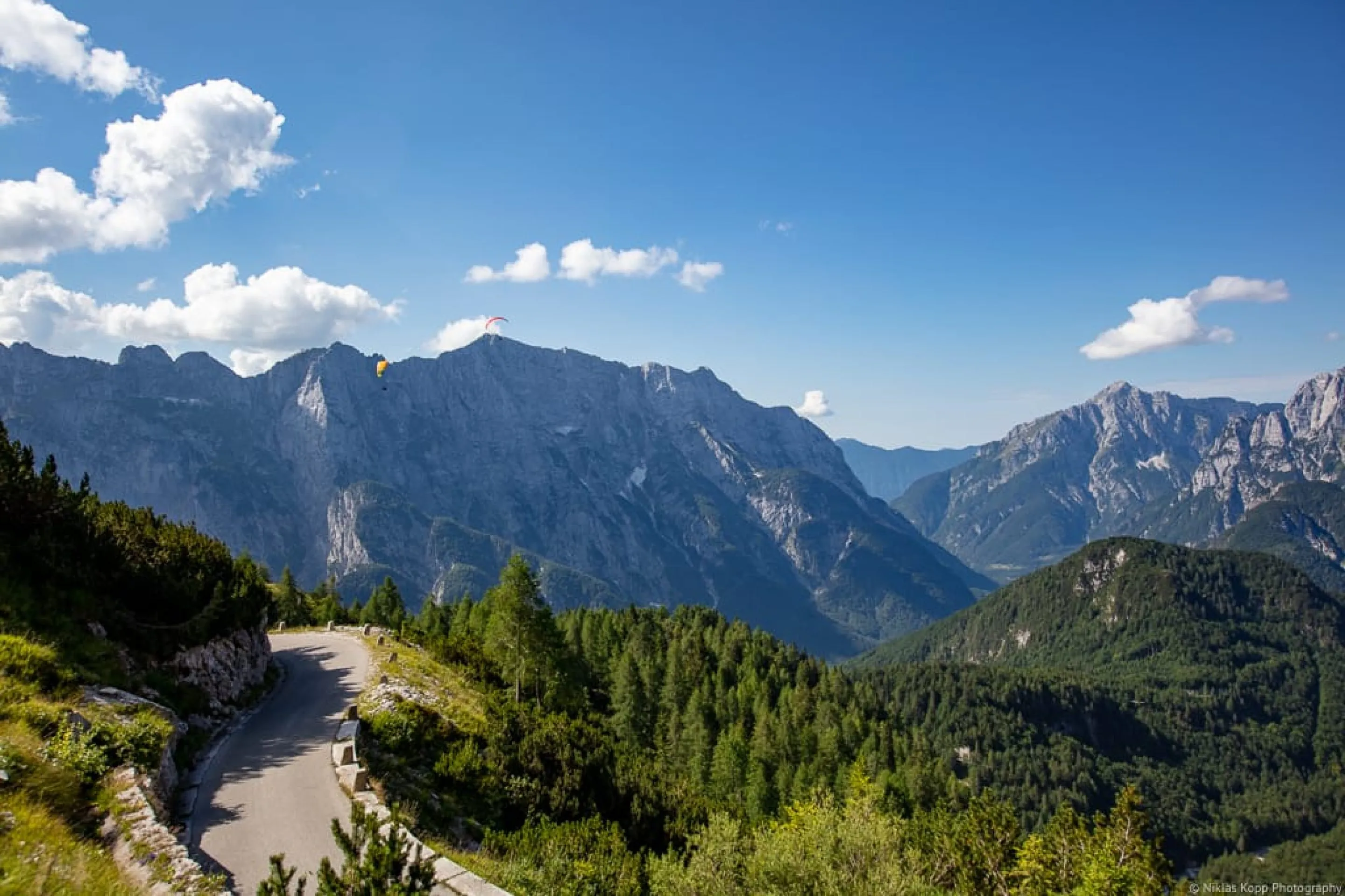 Eine schmale Bergstraße mit Gleitschirmfliegern am Himmel